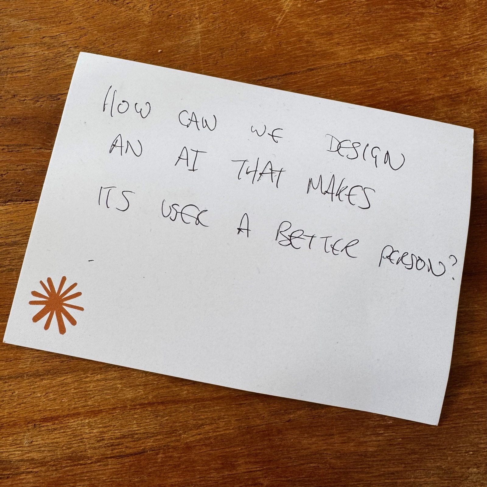 A handwritten index card reading 'How can we design an AI that makes its user a better person?', with an orange asterisk mark in the corner.